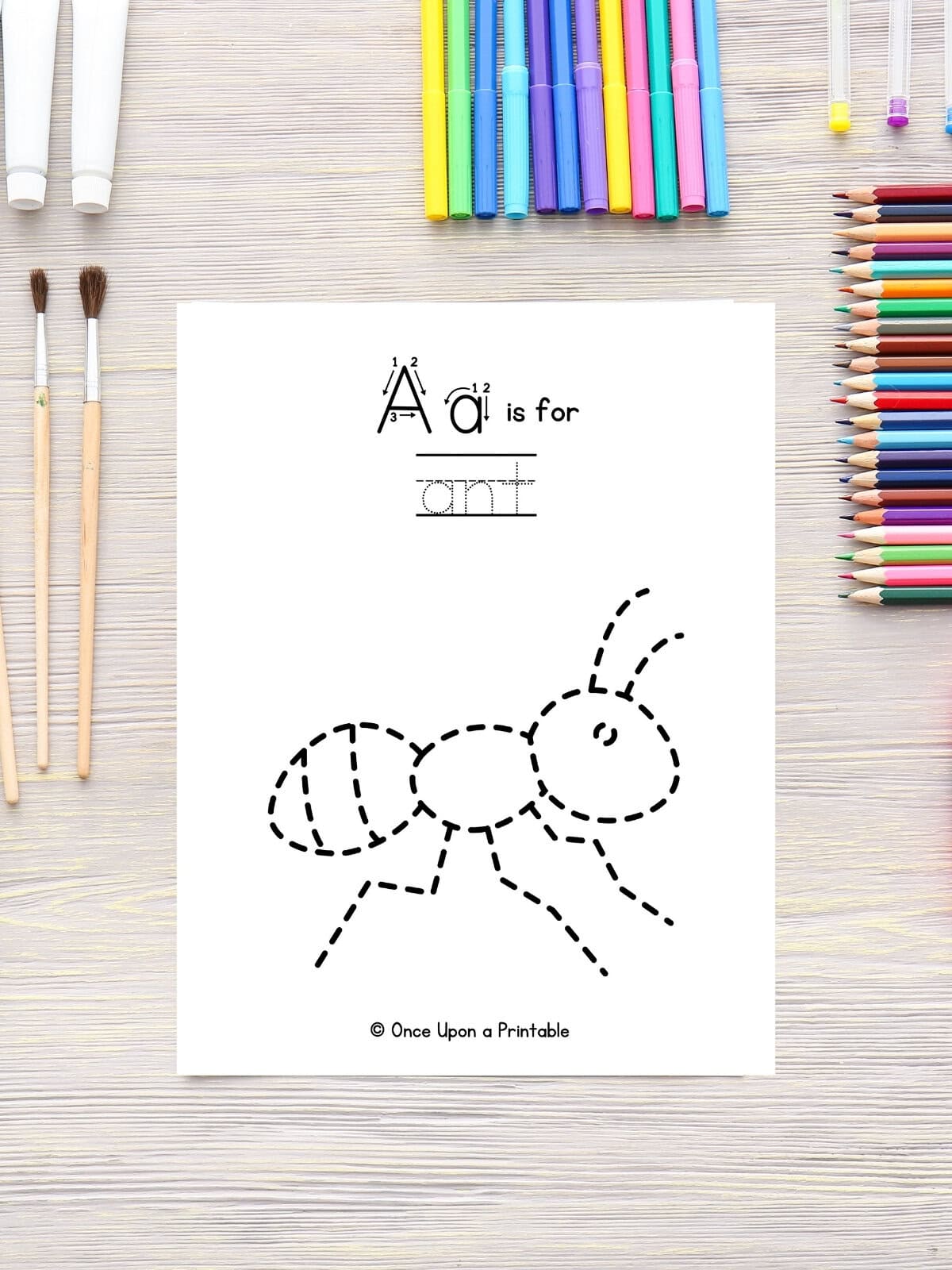 A is for ant, trace an ant and the word ant activity page laying on a table with coloring pencils, markers, and paint brushes.