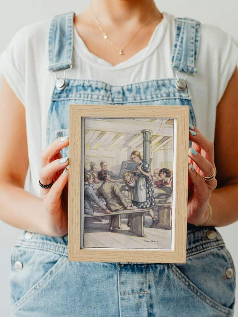Anne Shirley about to whack Gilbert Blythe on the head with her black board in class, illustration framed and held by a woman.