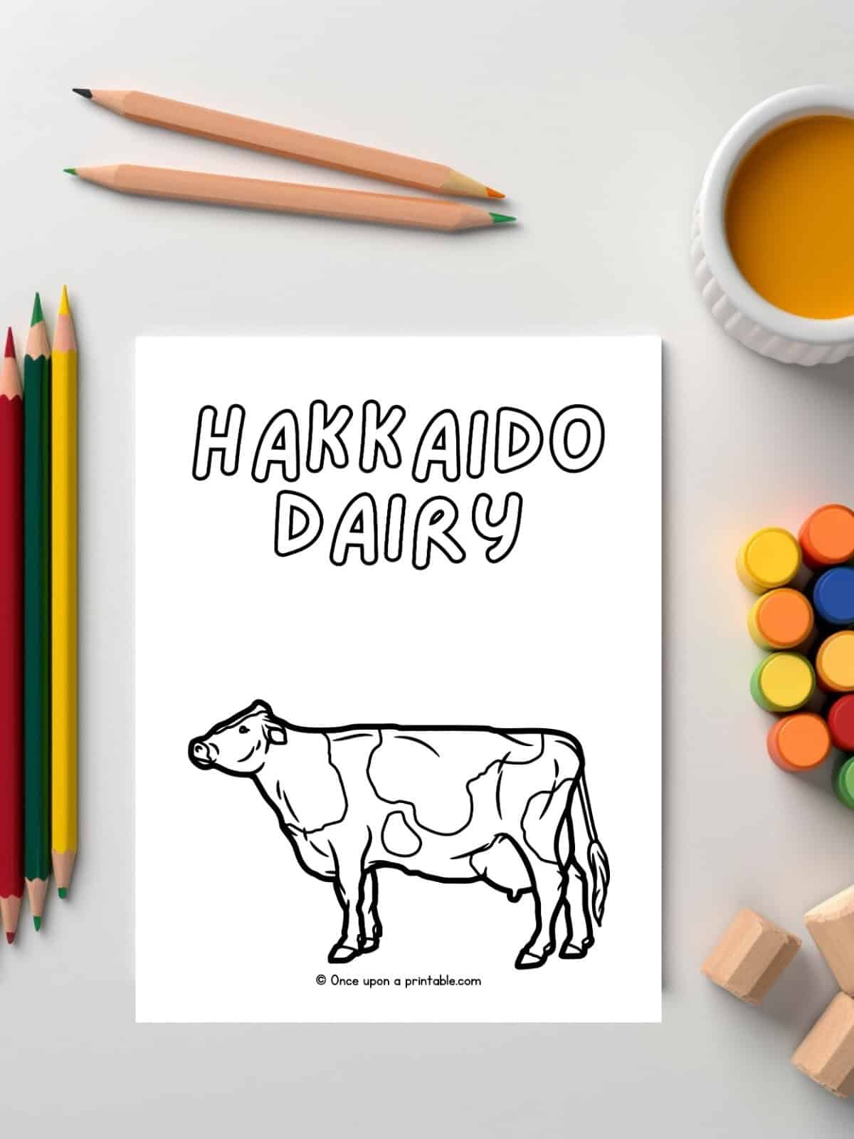 H is for Hakkaido Dairy cow coloring page laying on a white table with paints and pencils. 