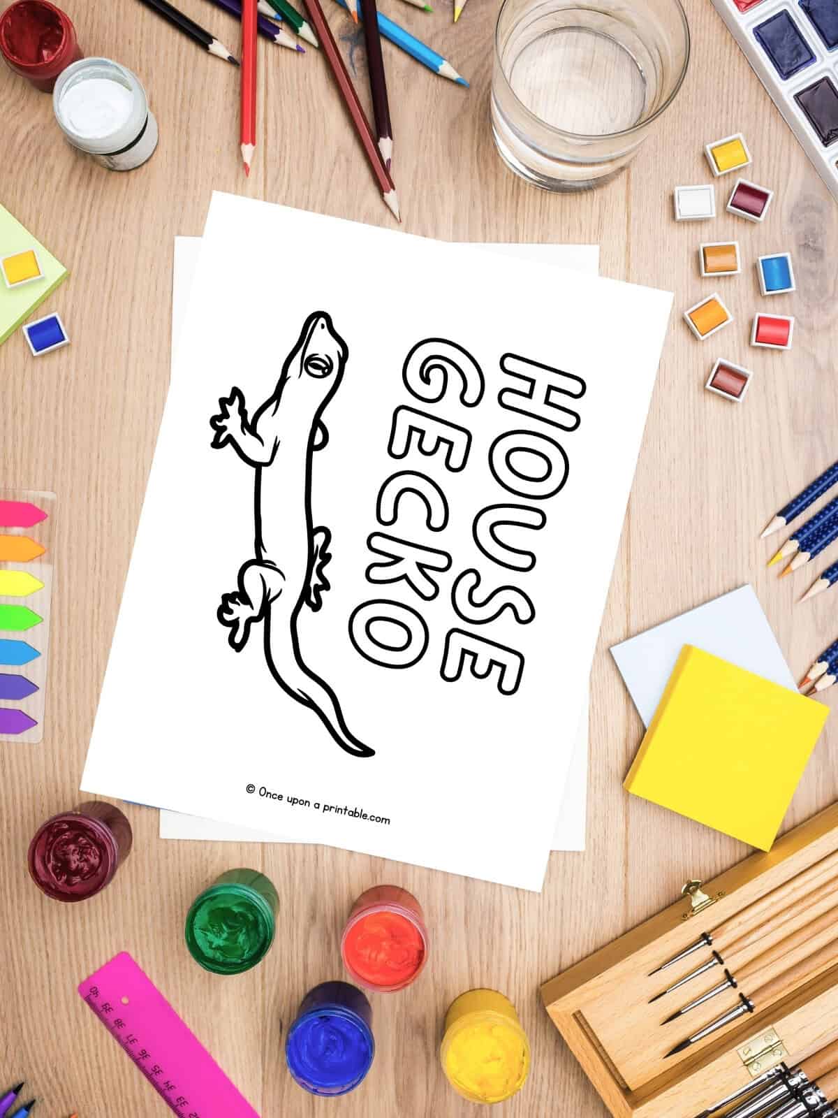 H is for House Gecko coloring page laying on a table with coloring supplies like crayons, pencils, and paint.