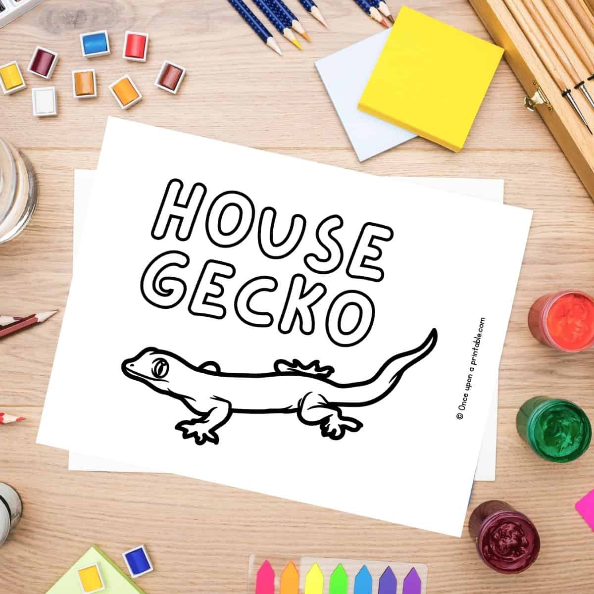 H is for House Gecko coloring page laying on a table with coloring supplies like crayons, pencils, and paint.