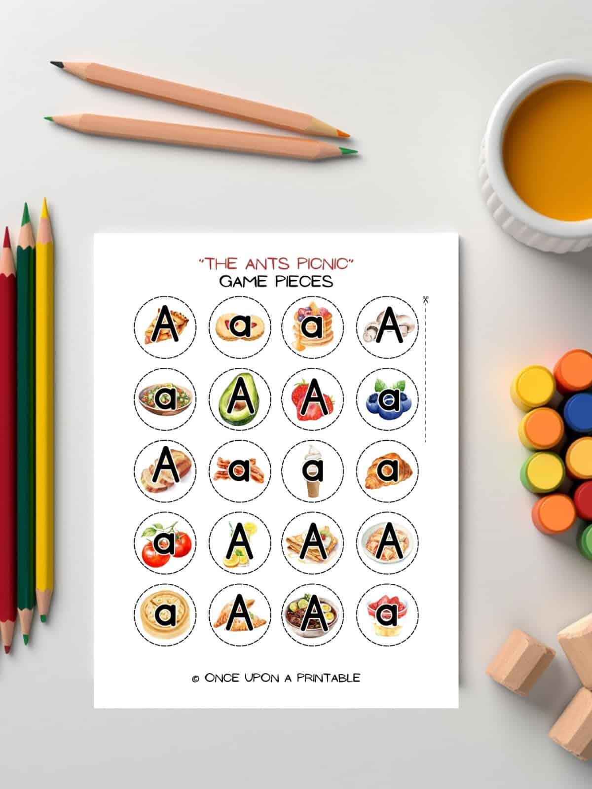 The Ants picnic game pieces with uppercase and lowercase A's laying on a table with paints and coloring pencils. 