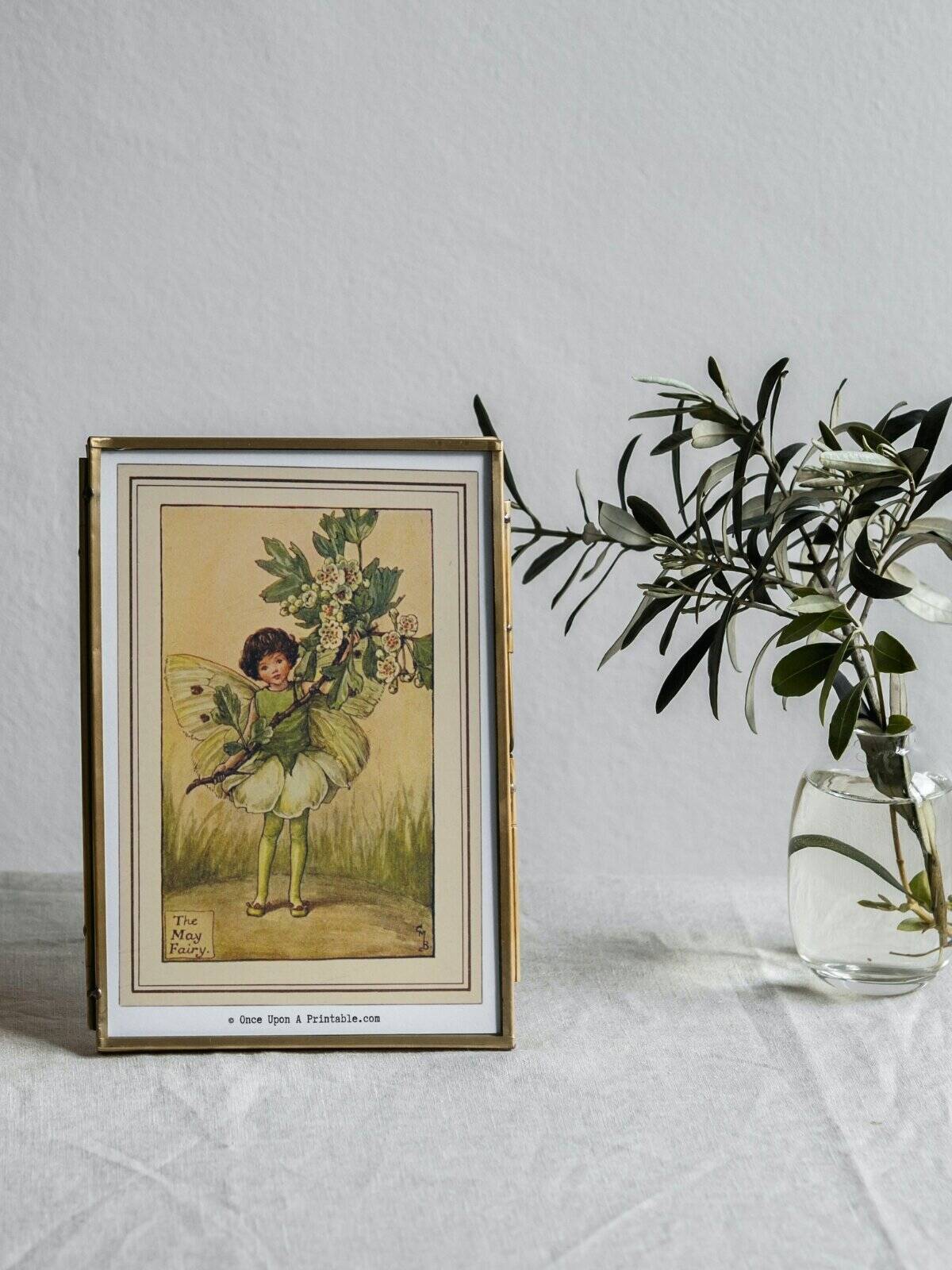 The May Flower fairy illustration framed and sitting next to a vase with a sprig.